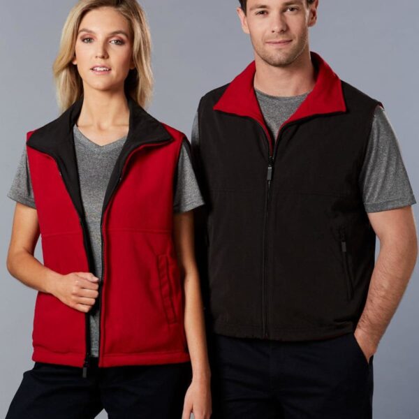 A woman and a man stand side by side, both wearing gray t-shirts under sleeveless zipped vests. The woman wears a red and black vest, while the man wears a black and red one. They both have dark pants and are smiling slightly.