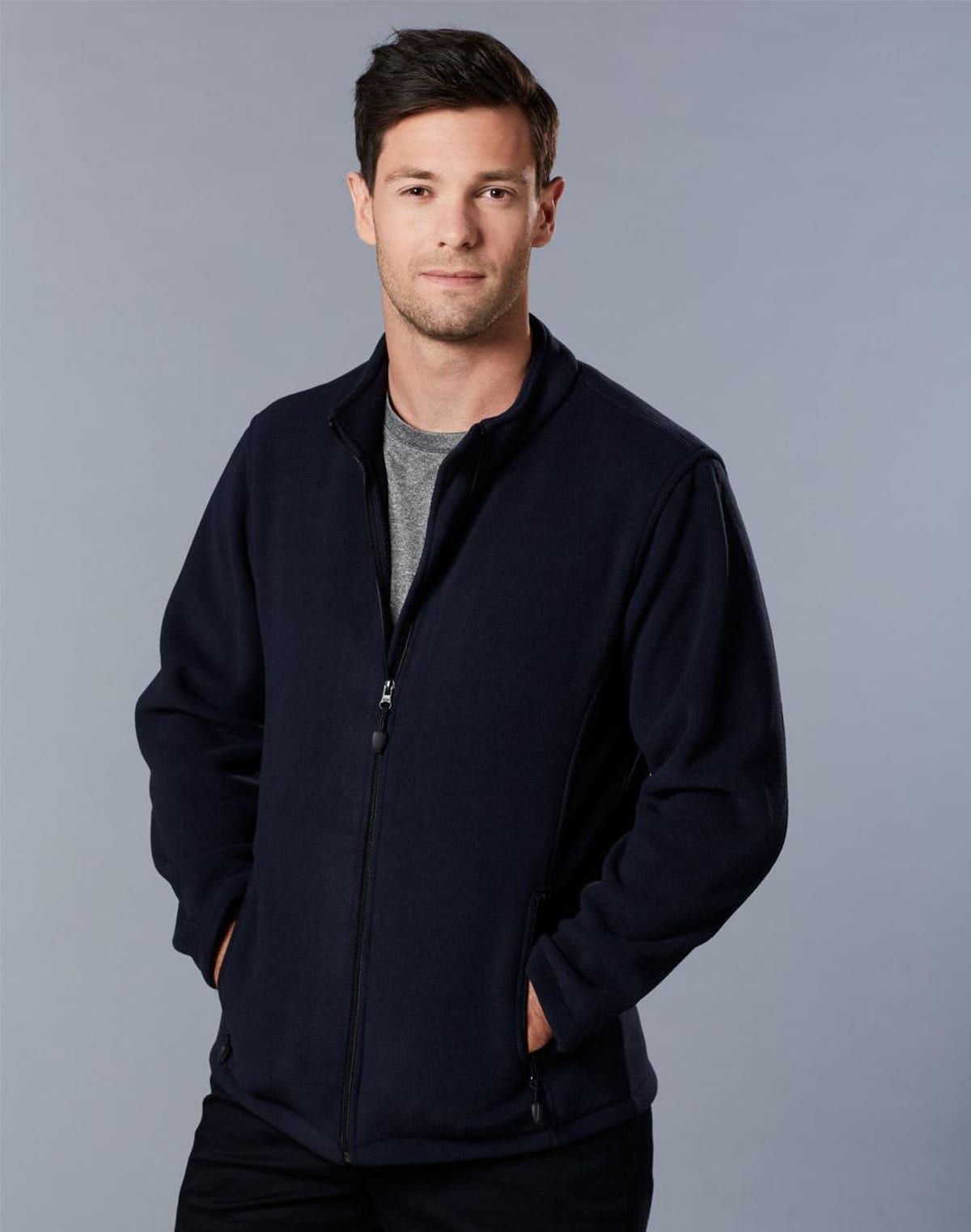 Man in a black zip-up fleece jacket stands against a gray background with hands in pockets, looking at the camera with a neutral expression. He has short dark hair and is wearing a grey t-shirt underneath.