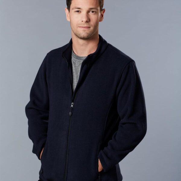 A man with short dark hair stands against a plain grey background, wearing a dark zip-up jacket over a grey t-shirt. His hands are in his jacket pockets, and he has a neutral expression.