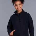 A smiling young person with curly hair wears a navy blue zip-up jacket with hands partly in the pockets. The background is a plain gray.