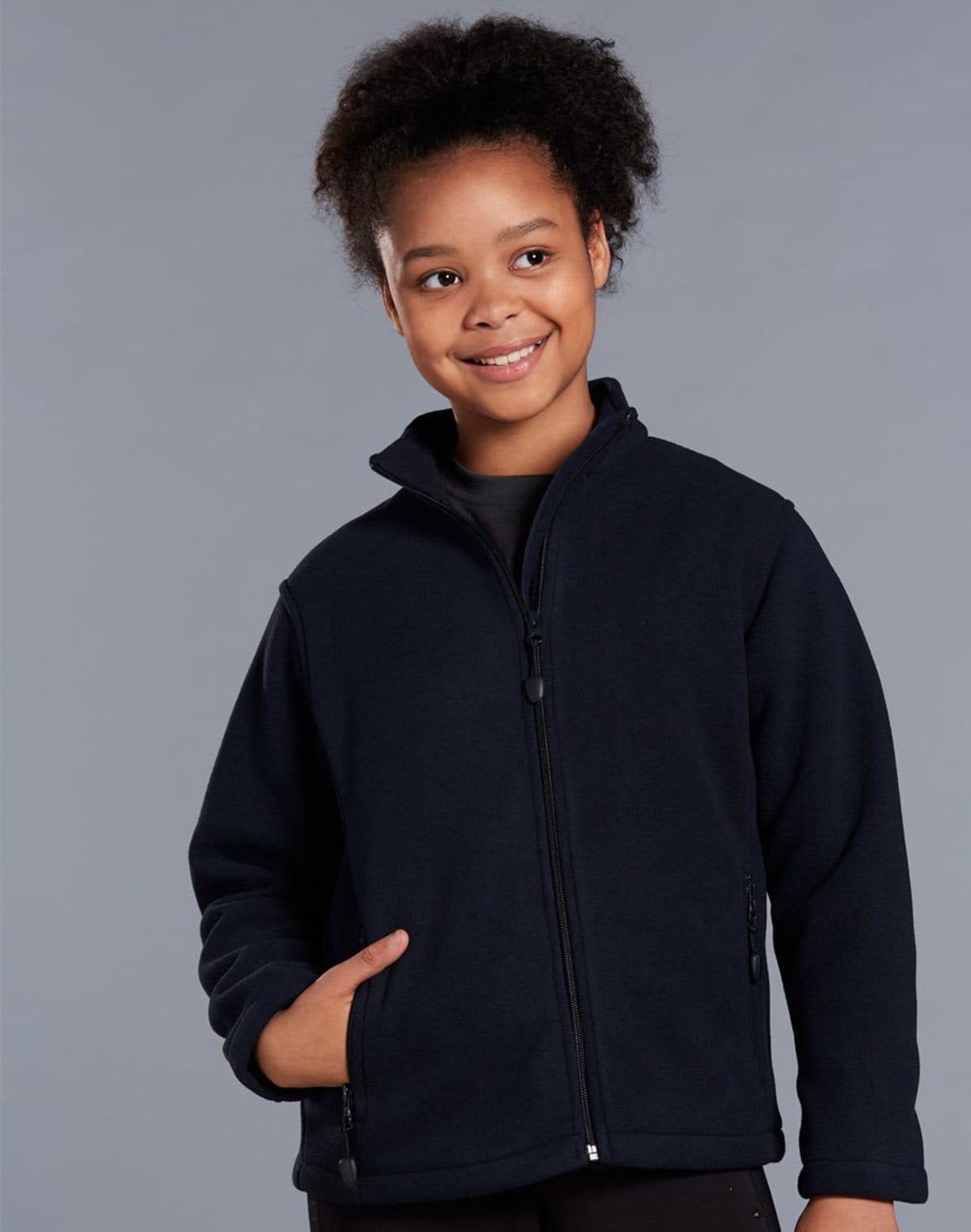 A smiling young person with curly hair wears a navy blue zip-up jacket with hands partly in the pockets. The background is a plain gray.