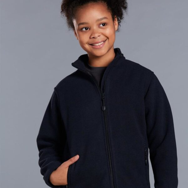 A smiling child with curly hair is wearing a navy zip-up jacket. They have one hand in their pocket and are standing against a gray background.