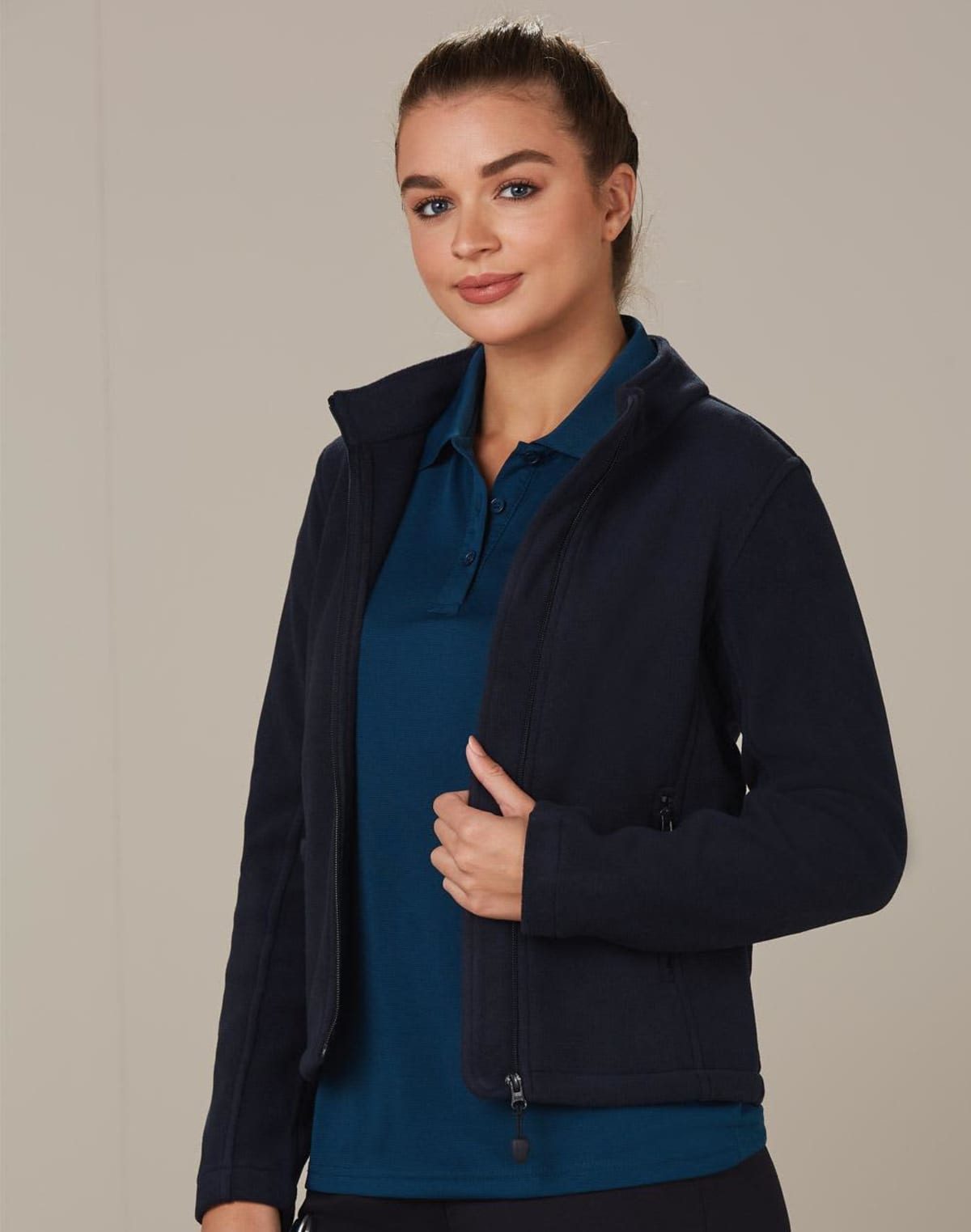 Person wearing a navy jacket over a teal polo shirt, standing against a plain background. They have their hair tied back and are smiling, holding the jacket with one hand.