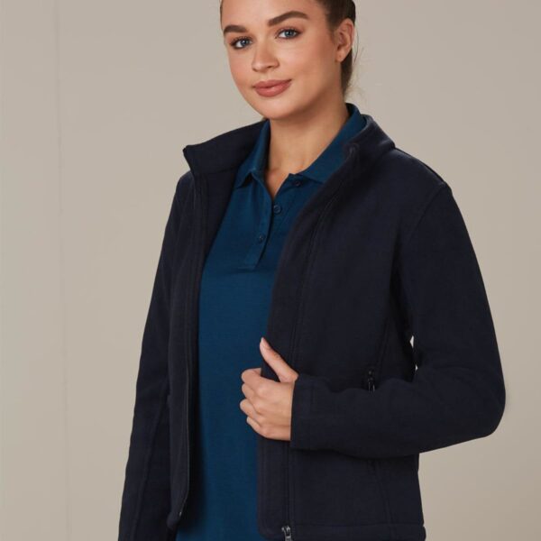 A person with hair in a bun is standing against a plain background. They are wearing a dark fleece jacket over a blue polo shirt, smiling slightly with one hand holding the jacket.