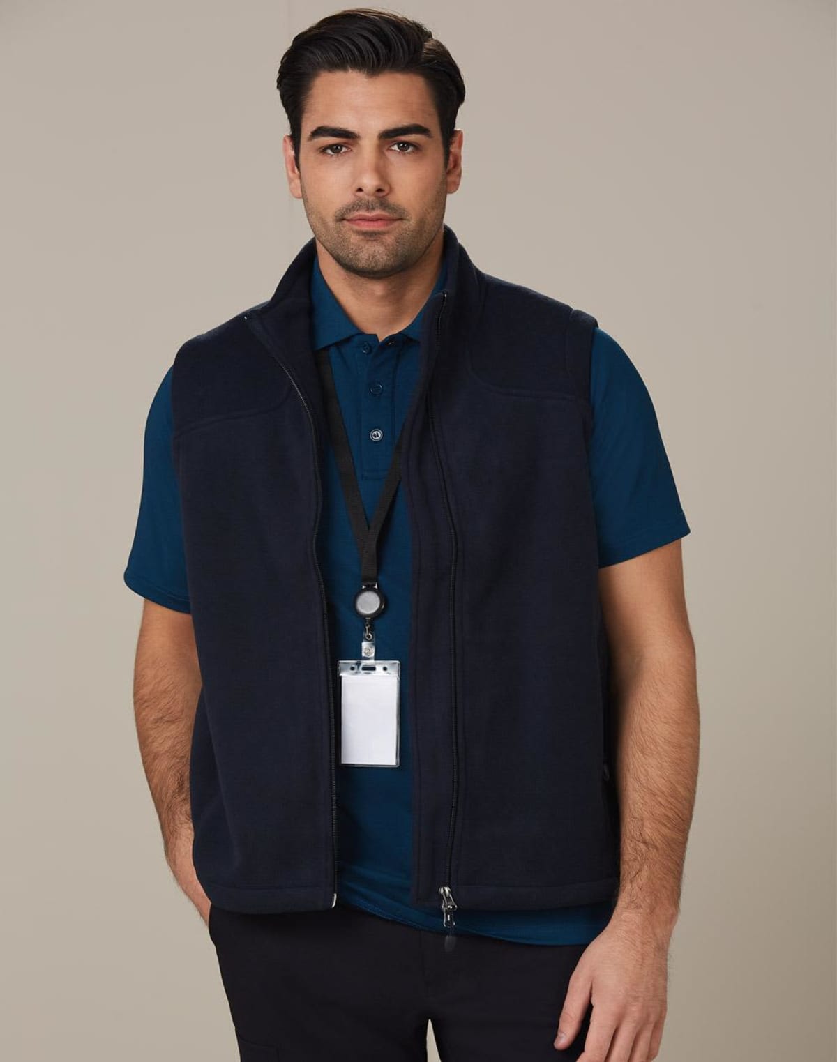 Man with dark hair and stubble wearing a dark blue fleece vest over a blue polo shirt. He has a lanyard with a blank ID badge around his neck and is standing against a plain background.
