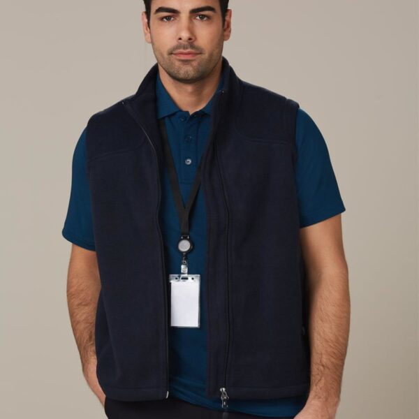 A man with dark hair is wearing a navy vest over a blue polo shirt. He has a lanyard with a badge around his neck. He stands against a plain, light brown background, looking directly at the camera.
