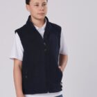 A young person stands against a plain background, wearing a dark blue zip-up vest over a white shirt and blue jeans. Their hands are in the vest pockets, and they are looking off to the side.