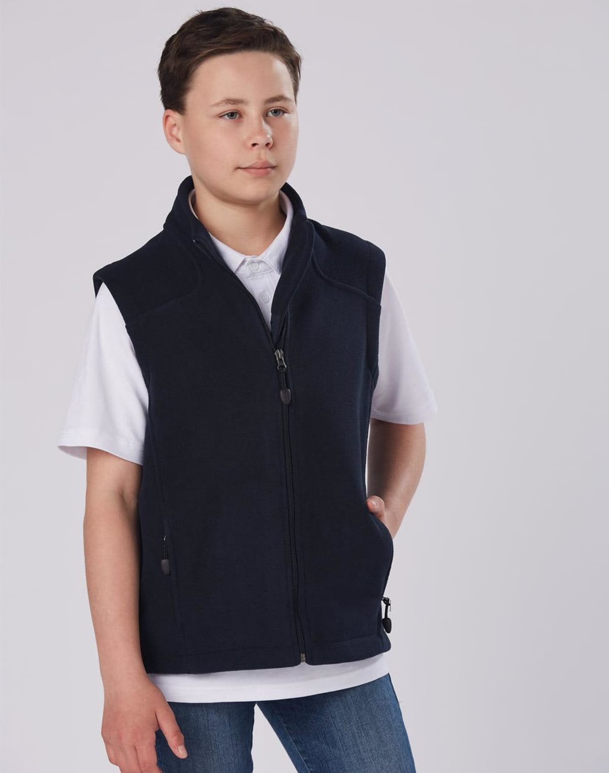 A young person stands against a plain background, wearing a dark blue zip-up vest over a white shirt and blue jeans. Their hands are in the vest pockets, and they are looking off to the side.
