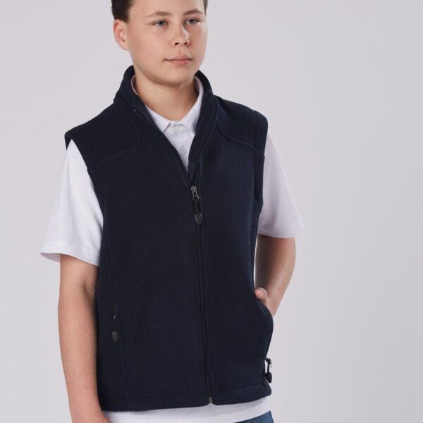 A young person with short hair is wearing a navy zip-up vest over a white polo shirt and jeans. The background is plain and light-colored.