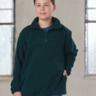 A person stands indoors wearing a dark green zip-up fleece pullover and blue jeans. They have short hair and are positioned against a backdrop with two large windows. One hand is in the pocket of the fleece.