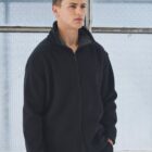 A person wearing a black zip-up jacket stands indoors, gazing to the side. They have short, dark hair and are in front of a large, square window with grid-patterned glass, which creates a soft, diffused light.