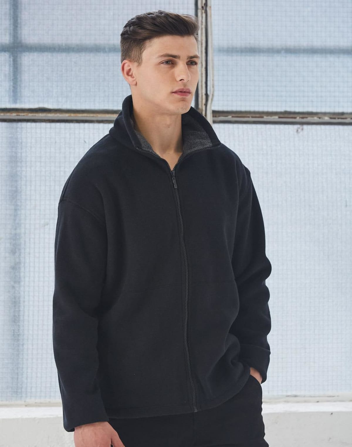 A person wearing a black zip-up jacket stands indoors, gazing to the side. They have short, dark hair and are in front of a large, square window with grid-patterned glass, which creates a soft, diffused light.