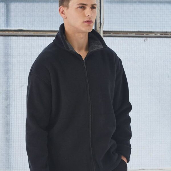 A man wearing a black zip-up jacket stands in front of a large window with a mesh-like covering. He has short, styled hair and is looking slightly to the side with a neutral expression. The setting appears to be indoors with natural light.
