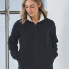 A person with shoulder-length hair wears a black fleece jacket with white lining, standing indoors near a window. They have their hands in the jackets pockets and are looking down, conveying a sense of casual comfort.