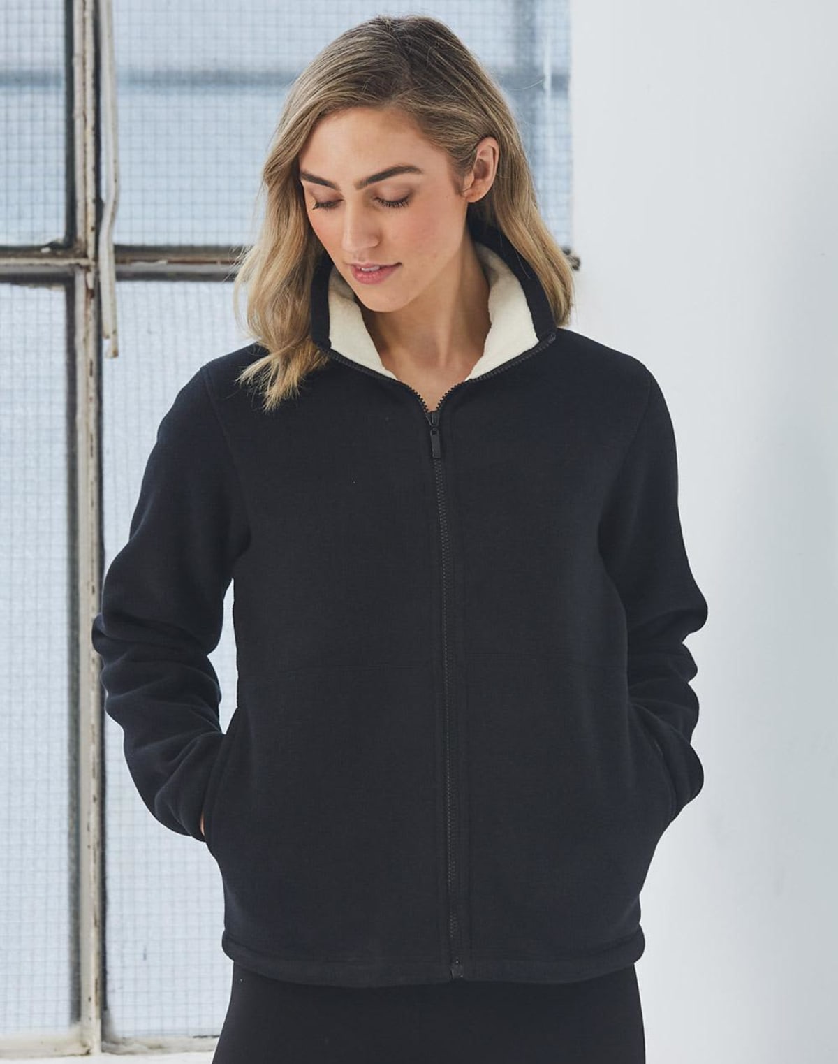 A person with shoulder-length hair wears a black fleece jacket with white lining, standing indoors near a window. They have their hands in the jackets pockets and are looking down, conveying a sense of casual comfort.