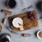 A cheese board with brie, a wedge cut out, surrounded by sliced bread and red grapes. A corkscrew, cheese knife, and wine bottle with glasses are nearby on a textured fabric surface.