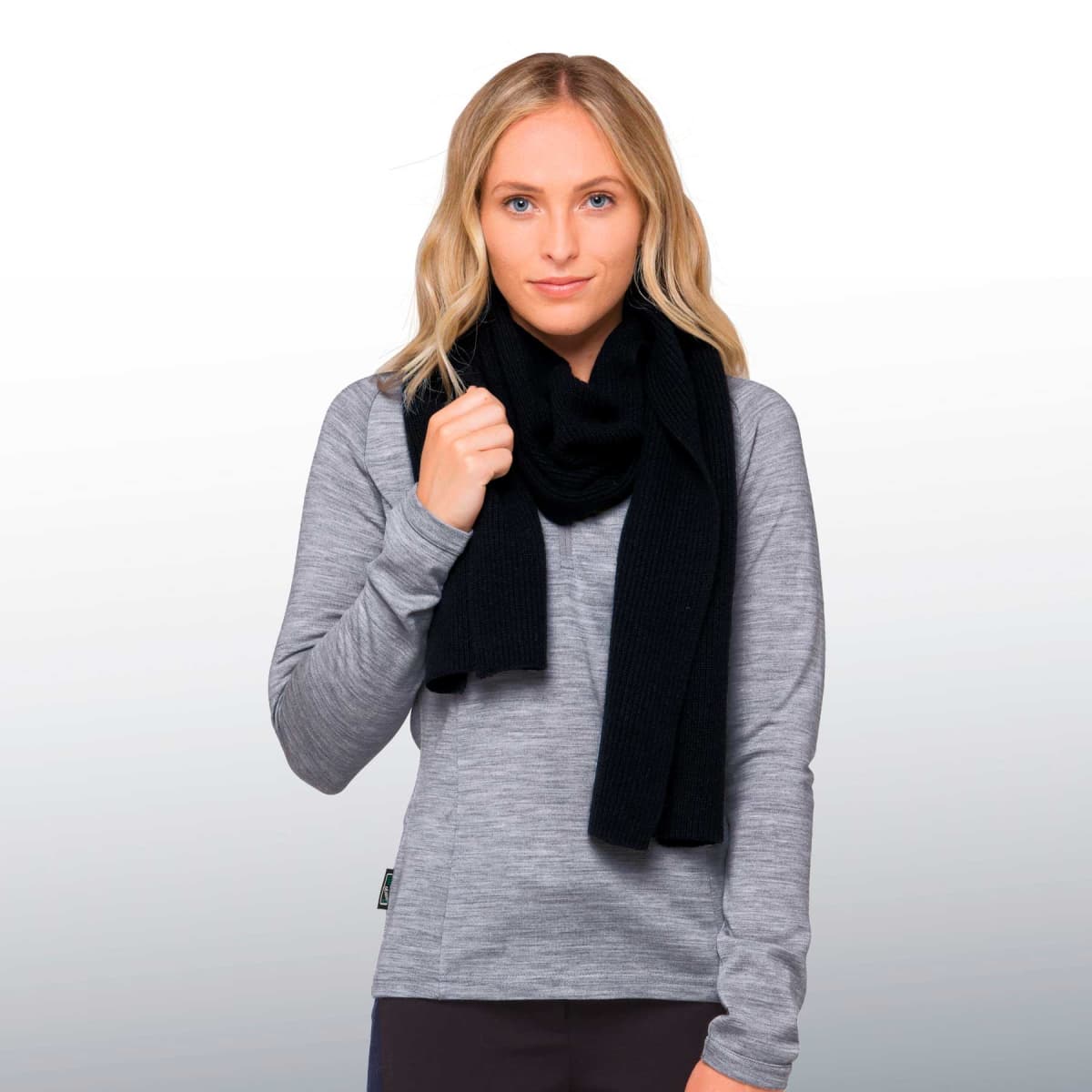 A person with blonde hair wearing a gray long-sleeve top and a black scarf stands against a light gray background. They are looking ahead, holding the scarf with one hand.