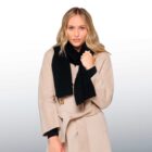 A person with long blonde hair wearing a beige coat and a black scarf, standing against a neutral background. The individual has a calm expression and holds one end of the scarf with one hand.