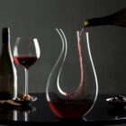 A bottle of red wine is being poured into a graceful, curvy glass decanter. A filled wine glass, a wine bottle, nuts, olives, and crackers are in the background on a dark table. The setting is elegant and moody.
