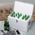 A white cooler filled with green beer bottles sits on a sandy beach. Three beer bottles are placed next to the cooler along with a bottle opener, black flip-flops, and sunglasses. Grassy dunes and calm sea are visible in the background.