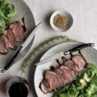 Two plates of sliced steak with greens, garnished with rosemary. A small bowl of seasoning sits nearby. A glass of red wine and two steak knives are also visible on the light-colored tablecloth.