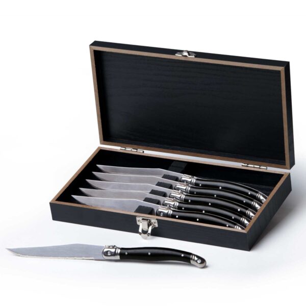 A set of six steak knives with black handles, neatly arranged in an open black wooden box. One knife is placed outside of the box. The box has a metal latch for secure closure.