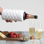 A hand pours champagne from a bottle wrapped in a textured white sleeve into two flutes. The table has a cheese board with grapes, strawberries, nuts, bread, and cheese. A cork lies nearby.