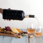 A hand pouring wine into two glasses on a wooden table, next to a spread of cheese, crackers, grapes, olives, apricots, and popcorn. A cork and a bottle are also visible on the table.