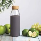 A glass bottle with a wooden cap and gray sleeve stands on a wooden table surrounded by green apples, limes—some sliced in half, and a bunch of green grapes. A hanging plant is visible in the background.
