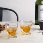 Two whiskey glasses filled with amber liquid sit on a table with a bottle of whiskey and a bowl of nuts nearby. A chair is partially visible in the background, and the table is covered with a light-colored cloth.