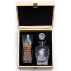 A wooden box with a black interior contains a whisky decanter set. Inside are a clear glass decanter with a stopper and two matching glass tumblers, alongside a labeled paper insert indicating The Islay Whisky Decanter Set.