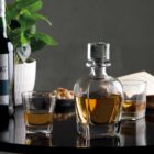 A stylish glass decanter filled with amber liquid is placed on a round black table, accompanied by two glasses, a plant, an open book, and a wine bottle. The setting is elegant and inviting.