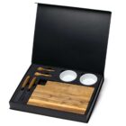 A black gift box containing a wooden cutting board, two small white bowls, two cheese knives, and two small wooden serving spoons. The items are neatly arranged within custom black compartments.