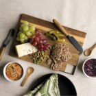 A wooden board with grapes, blue cheese, pickles, pistachios, and seed crackers. Two butter knives and a napkin are nearby. A small bowl of dip and another of red jam are on the table. All items are arranged on a light linen tablecloth.