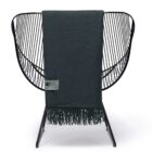 A black wire-frame chair with a dark green throw blanket draped over it. The blanket has fringe at the bottom and features a small label with text near the top. The chair has an open, modern design.