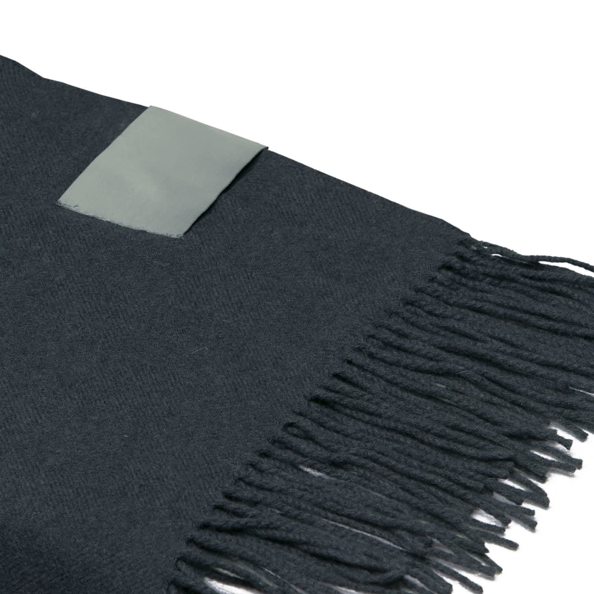 A close-up of a dark-colored fabric with fringed edges. A small rectangular label is stitched near the top corner, contrasting slightly with the fabric.