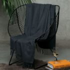 A modern black metal chair with a dark gray throw draped over it sits in a minimalist room with a concrete wall. A stack of books and a glass of white wine are on the floor beside the chair. A leafy plant is partially visible in the background.
