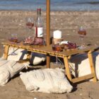 A wooden beach table with wine glasses, a bottle, a candle, grapes, and charcuterie is set on sand with decorative cushions. The backdrop features the ocean and a serene beach setting.