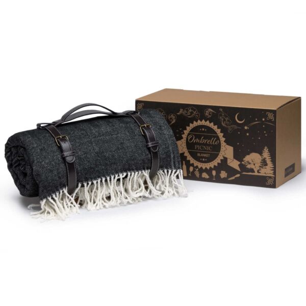 A rolled-up dark gray picnic blanket with white fringes, secured with black leather straps, is placed next to a decorative cardboard box. The box has artistic illustrations and the text Ombrello Picnic Blanket.