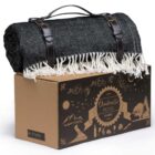 A rolled-up black and white herringbone picnic blanket with fringes, secured with leather straps, sits atop a decorative cardboard box. The box features outdoor-themed illustrations and the text Ombrello Picnic Blanket.