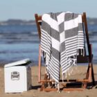 A wooden beach chair with a black and white striped towel draped over it, set on a sandy beach. Next to the chair is a white cooler with a partially open lid. The ocean and a distant shoreline are visible in the background.