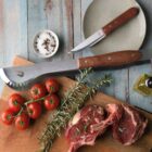 A wooden table with fresh tomatoes, a salt dish, rosemary sprigs, raw seasoned steaks, olive oil, a large knife, and a small knife arranged on a cutting board. A plate and additional cutlery are nearby.