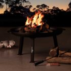 A fire pit with a bright, burning fire is set on a patio at dusk. Logs and fire pokers are nearby. Two cups and a pile of marshmallows are on the ground, suggesting a cozy outdoor gathering. Trees are silhouetted against the evening sky.