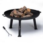 A black metal fire pit with four sturdy legs, filled with stacked firewood pieces. A fire poker rests on the ground next to it. The setup is on a plain white background, showcasing the fire pits design and accessories.