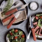 A table setting with steak knives on a napkin, a bowl of salt, and two plates. One plate holds sliced steak and salad with cherry tomatoes and greens, while the other contains a mixed salad with tomatoes and feta cheese. Cutlery is placed nearby.