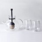A clear glass teapot with a black handle and a mesh infuser inside is placed on a white surface. Next to it are two clear, stemless glass cups. The background is a light gray wall.