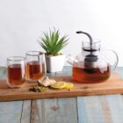 A clear glass teapot with tea, two filled glasses, a small potted plant, ginger, lemon slices, and dried chamomile flowers rest on a wooden board on a blue wooden table.