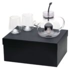 A clear glass teapot with a metal infuser, accompanied by two glass cups, all placed on a black rectangular box. The teapot has a handle and a curved spout. The glasses are cylindrical with a slight curvature.