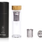 A glass tea infuser with a bamboo lid next to a cylindrical black packaging container. Two metal infuser components are displayed to the side. The container has po di fame and little hungry printed on it.