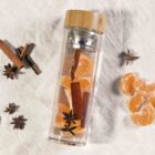 A glass bottle with a bamboo lid contains water, mandarin slices, cinnamon sticks, and star anise. Next to it, more mandarin slices and star anise pieces are scattered on a textured beige cloth.